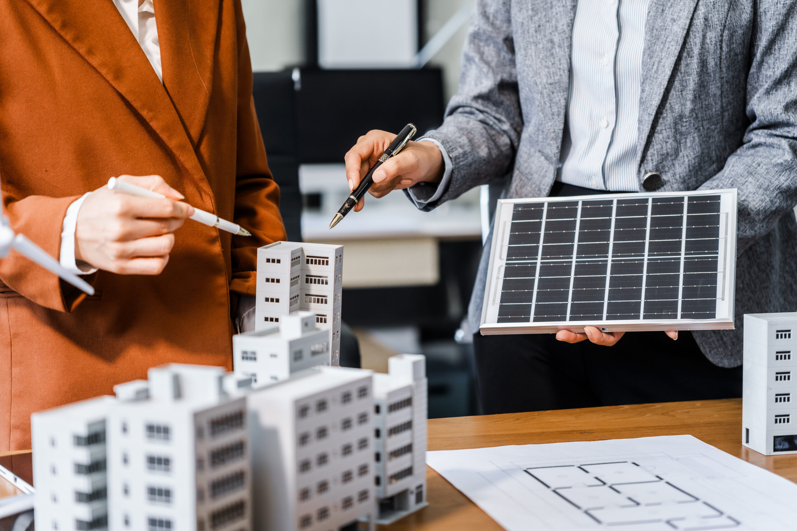 Two business women are talking. One is holding a solar panel while the other is pointing a model of a commercial building